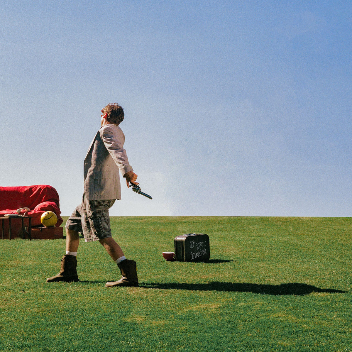 A man in a light-colored blazer, shorts, and boots stands on a grassy field holding a pistol, with a red sofa, a yellow ball, and a black suitcase under a clear blue sky.