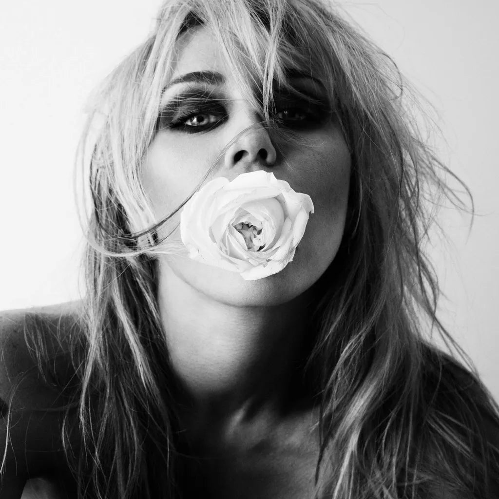 Black and white portrait of a woman with dramatic dark eye makeup holding a white rose to her lips, shown against a plain light background.