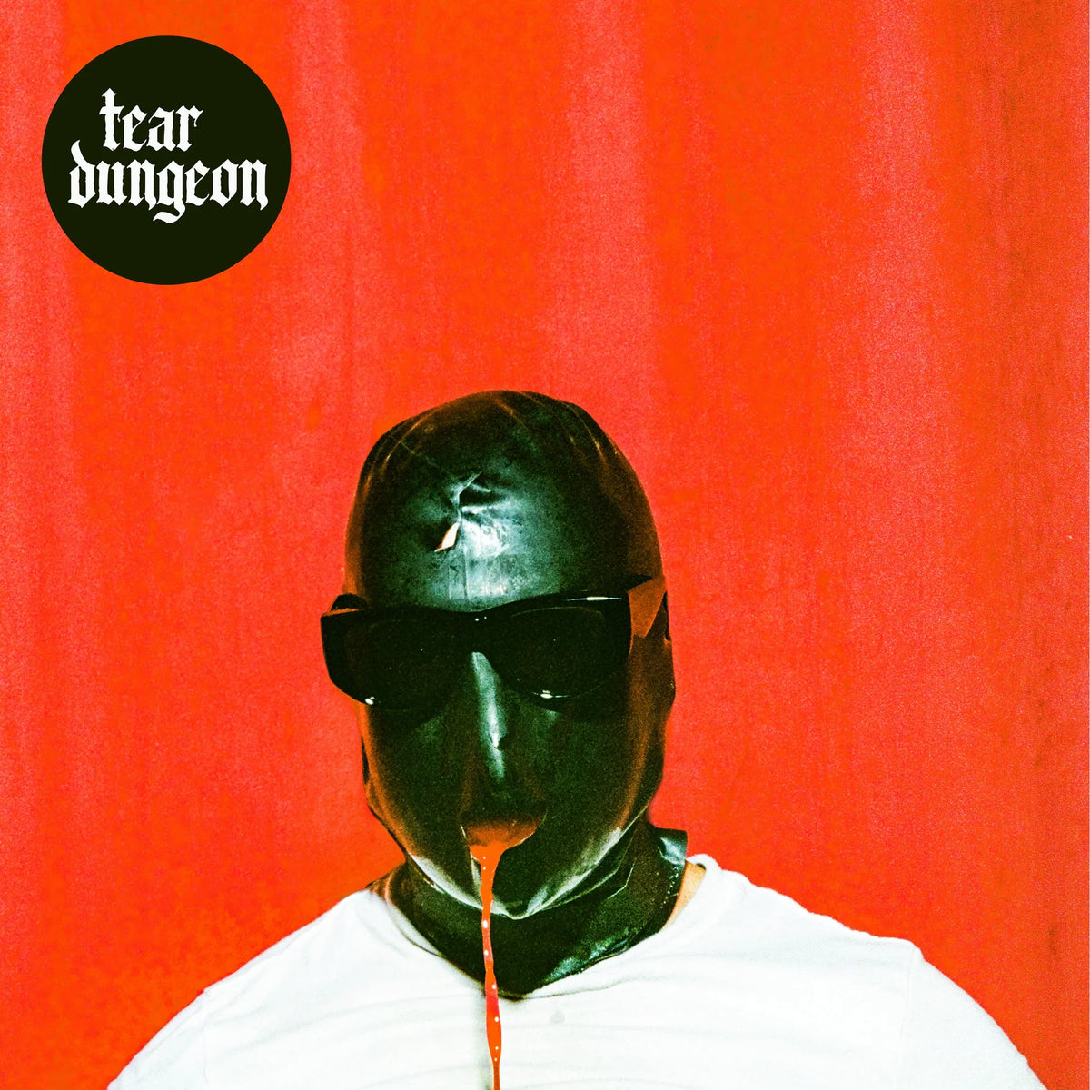 Album cover art for 'Tear Dungeon' featuring a person in a glossy black latex mask and sunglasses against a bright red background, with a black circular logo in the top-left corner.