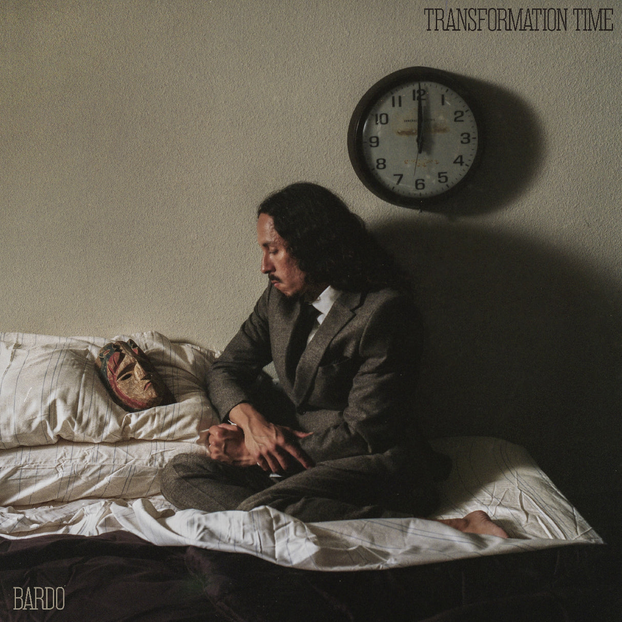 Man in a suit sitting on a bed with a mask next to him, clock on the wall, and text 'Transformation Time' above.