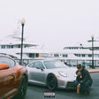 Person squatting next to a white sports car with a large ship in the background
