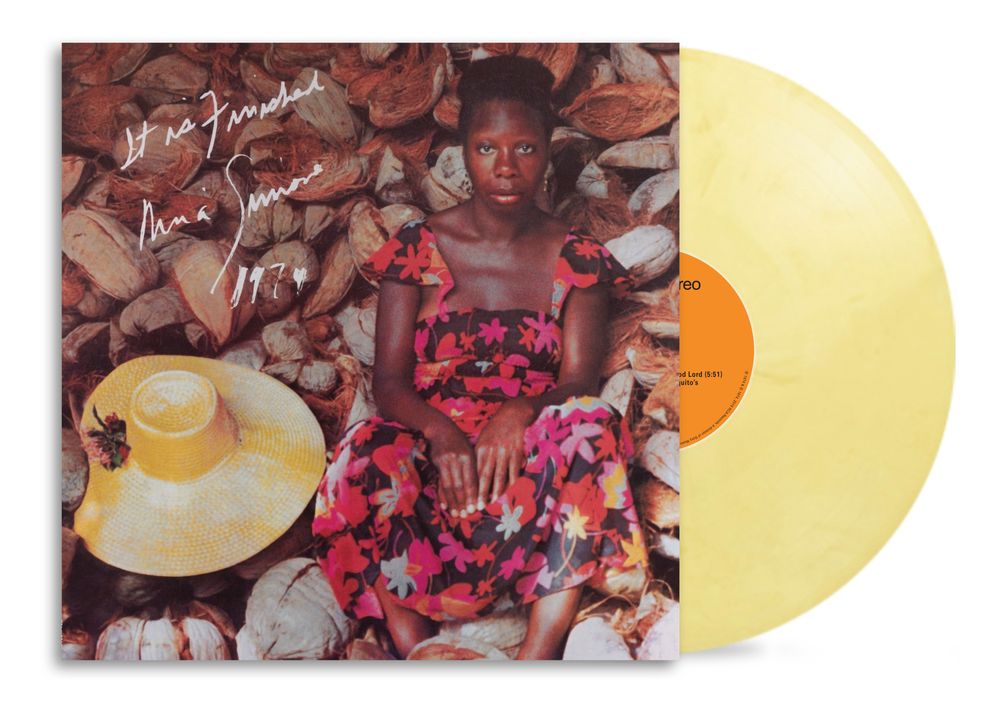 A vinyl record album shown with its cover partially pulled out, revealing a pale yellow translucent record. The cover features a portrait of a seated woman surrounded by brown coconuts, with a yellow hat in the lower left and white handwritten text near the upper left.