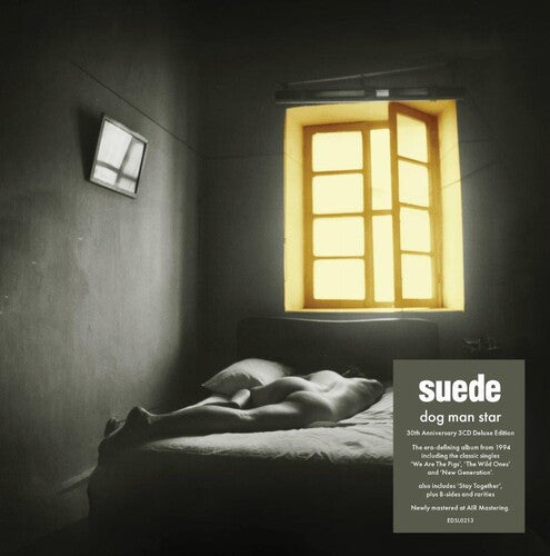 The image depicts the cover of the album 'Dog Man Star' by the band Suede, celebrating its 30th anniversary. The cover features a black and white photograph of a person lying on a bed in a dark room, with a yellow window frame illuminating the space.