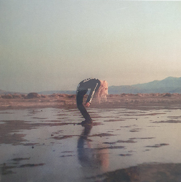 An album cover featuring a person crouched down on a wet surface with a landscape in the background.