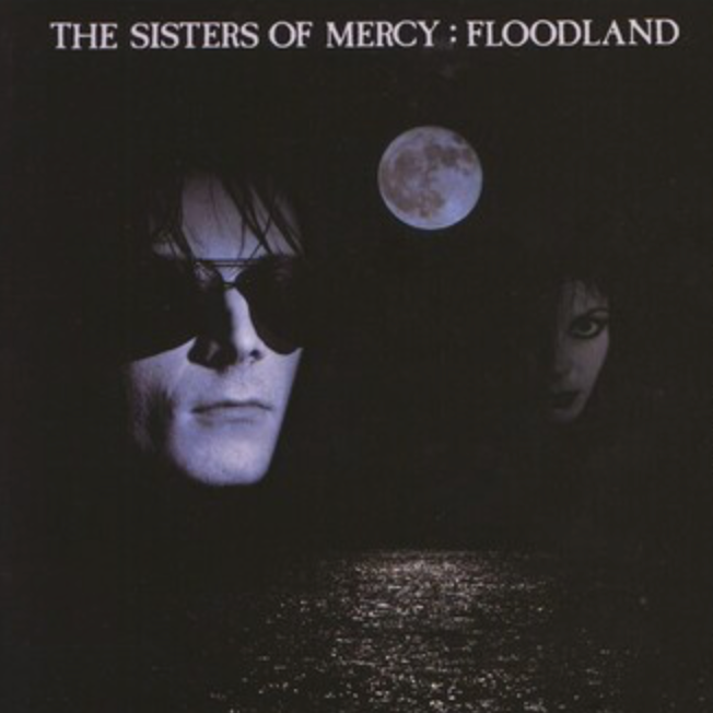 The image features the album cover for "The Sisters Of Mercy - Floodland LP," showcasing an eerie and atmospheric scene. The cover depicts a dark, foreboding nightscape with a large, glowering moon in the background and the silhouette of a man and a woman in the foreground.