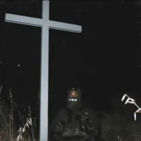 A person wearing a mask sitting in front of a cross in a dark environment, possibly as a symbolic or artistic representation related to the album's themes.