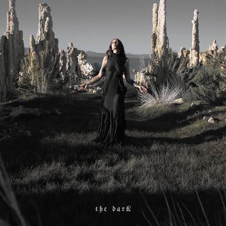 A woman in a long black dress stands in a dramatic outdoor landscape with tall rock spires and dry grass. The photo is mostly grayscale, with the title 'the dark' at the bottom.