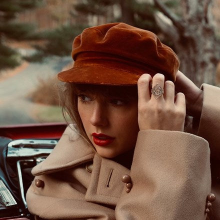 Taylor Swift wearing a red cap and beige coat, sitting in a car, promoting her album 'Red: Taylor's Version'.