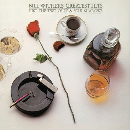 Album cover for Bill Withers' Greatest Hits featuring a rose, a cup of coffee, a whiskey decanter, and a ashtray with a cigarette on it, arranged on a table.