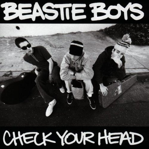 The image features the cover of the album "Check Your Head" by the Beastie Boys. The album cover displays a black and white photograph of the three members of the band, sitting closely together against a wall, with a suitcase-style guitar case in front of them.