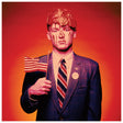 A man in a dark patterned suit and striped tie stands against a red-orange gradient background, holding a small American flag. His face is painted to resemble Donald Trump, with blond hair and red streaks.