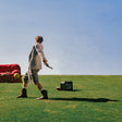 A man in a light-colored blazer, shorts, and boots stands on a grassy field holding a pistol, with a red sofa, a yellow ball, and a black suitcase under a clear blue sky.