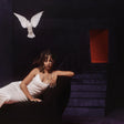 A woman in a white sleeveless dress reclines on a dark chair in a dim interior, with a white dove flying above and a glowing red-orange rectangular light on the stairs behind her.