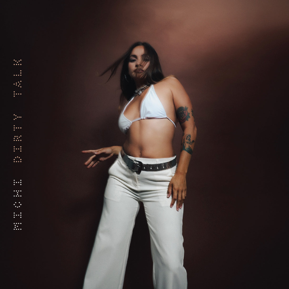 An album cover featuring a Latinx artist in a white crop top and pants, posing against a brown background.