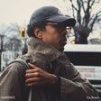 Man wearing a cap, glasses, and plaid scarf on a city street with 'ODDISEE' branding.