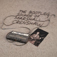 Cassette tape and photo with 'The Bootleg Sounds of Marshall Crenshaw 1984-1987' text on a textured surface.