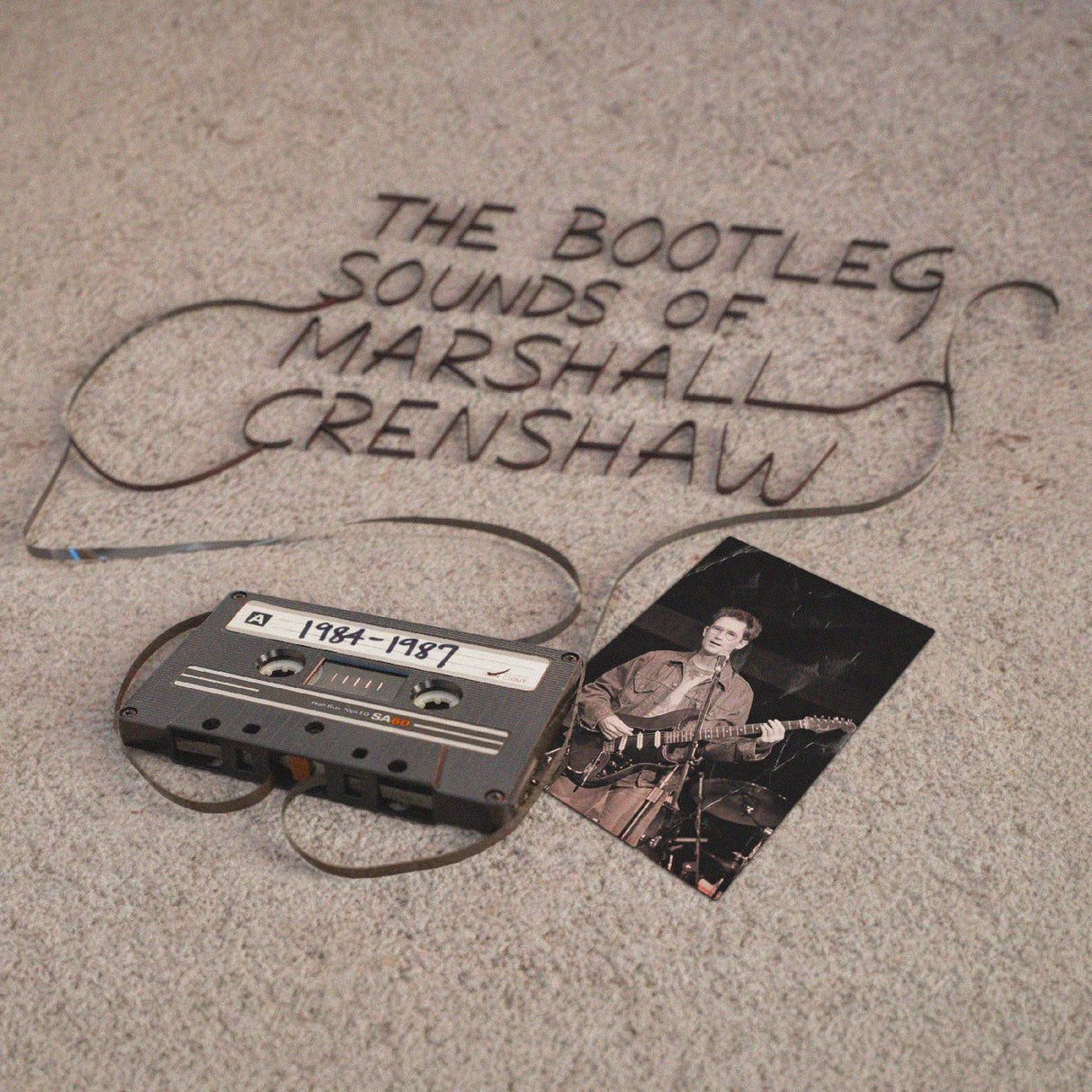 Cassette tape and photo with 'The Bootleg Sounds of Marshall Crenshaw 1984-1987' text on a textured surface.