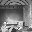 Two people wearing masks sitting on a couch in a room with curtains and a light switch.