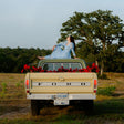 A person sitting on the back of a vintage Ford truck, with a guitar and a backdrop of a forest and a setting sun.