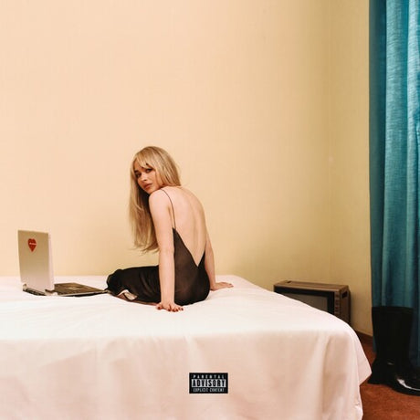 A person sitting on a bed with a laptop and a heart-shaped pendant, looking at the camera.