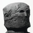 A dark charcoal-colored sculptural bust of a face with highly textured, weathered details, set against a plain white background.