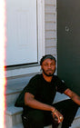A person sitting on a step outside a house, wearing a black t-shirt and shorts, with a relaxed posture.