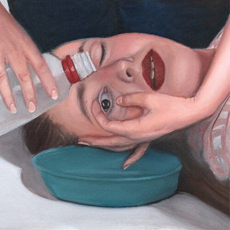 Person lying on a white surface with a teal bowl under their head, holding a bottle above their face.