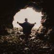 Person standing in a dark cave with arms outstretched, facing a bright light at the cave's entrance.