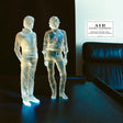 Clear plastic figures of two people on a dark surface with 'AIR' album cover in the background.