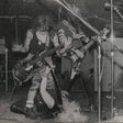 Two musicians playing electric guitars on stage with a microphone stand in the foreground.