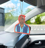 A die-cut air freshener or car fragrance hanging from the rearview mirror inside a car. The product features a stylized portrait of Tupac Shakur with a red outline, blue shirt, and dark vest, set against a blurred car interior and outdoor background.