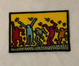 A rectangular embroidered patch featuring a yellow background with stylized dancing figures in red, light blue, olive green, magenta, and orange, outlined in black.