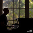 Silhouette of a person playing a piano in front of a large window with greenery outside
