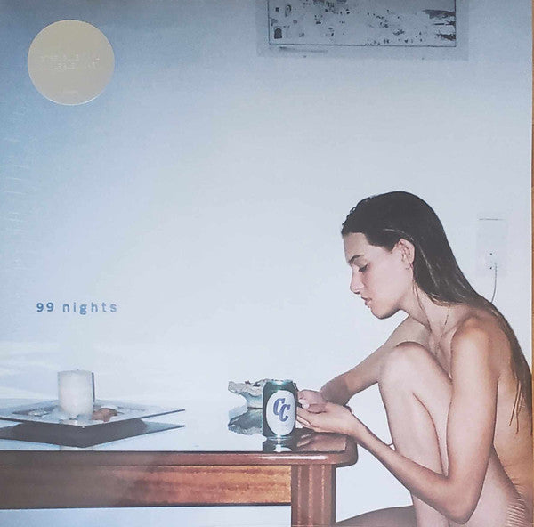 A woman sitting at a table with a record labeled '99 nights' in front of her. The setting appears to be a studio environment with a plain background.