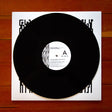 A black vinyl record partially pulled from a white square record sleeve with bold black geometric border graphics, placed on a brown wooden surface. The center label shows artist text including 'Troy Gatty' and 'Amenra'.