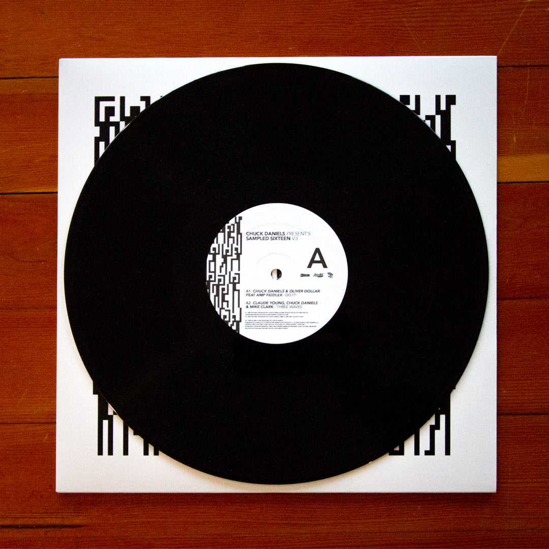 A black vinyl record partially pulled from a white square record sleeve with bold black geometric border graphics, placed on a brown wooden surface. The center label shows artist text including 'Troy Gatty' and 'Amenra'.