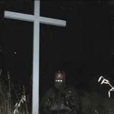 A person wearing a mask sitting in front of a cross in a dark environment, possibly as a symbolic or artistic representation related to the album's themes.