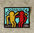 An embroidered patch featuring two stylized dancing figures in yellow and red on a light blue background, with black outlines and a black border, placed on a gray textured fabric surface.