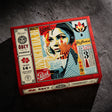 A boxed jigsaw puzzle package photographed on a dark textured surface. The box is red and cream with illustrated collage-style artwork featuring a woman's face, fan motifs, and text including 'Obey', 'Quality', '1000 pieces', and 'For ages 14+'.