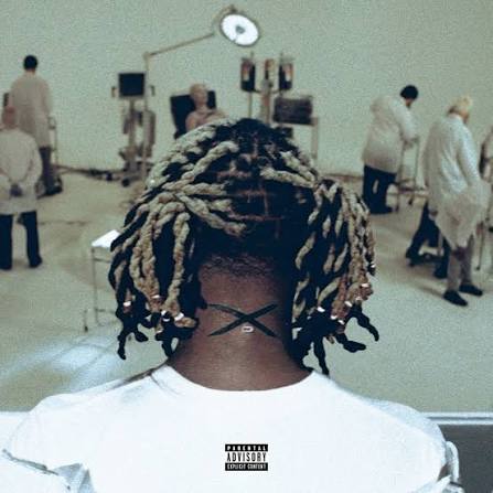A person viewed from behind wearing a white T-shirt, with braided dreadlocks and a black X tattoo on the back of the neck. The background shows a sterile, clinical room with several people in white coats and medical equipment.