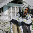 A person wearing a patterned headscarf covering the lower half of their face, standing in front of a white fence and a house with a gated entrance.