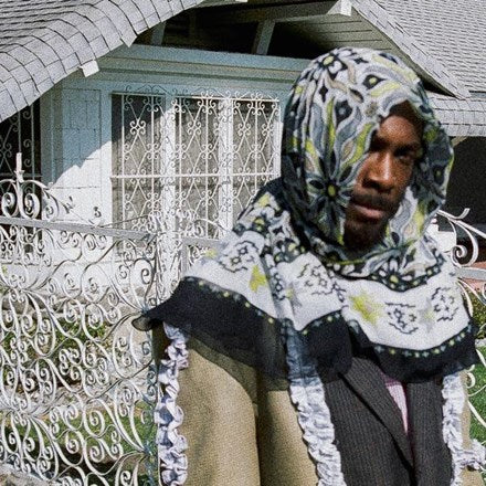 A person wearing a patterned headscarf covering the lower half of their face, standing in front of a white fence and a house with a gated entrance.