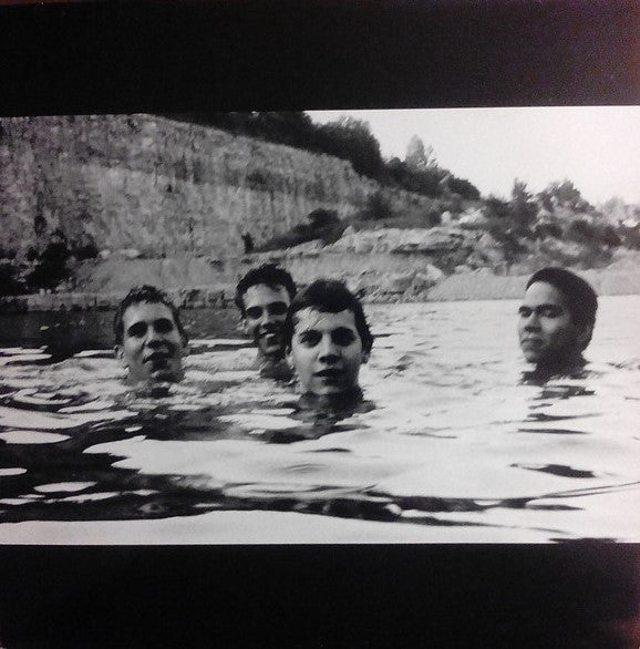 A black and white photo of four people standing in a body of water, likely a lake or a pond, with a rocky background.