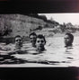 A black and white photo of four people standing in a body of water, likely a lake or a pond, with a rocky background.