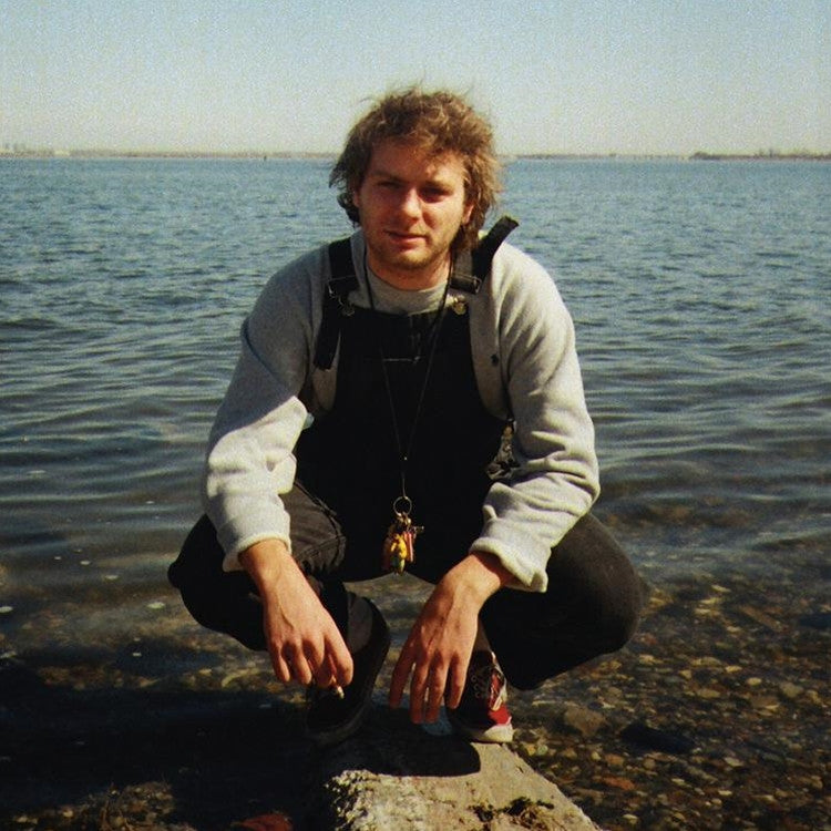 A photo of Mac DeMarco sitting by a body of water, wearing a grey sweater and black pants, with a camera around his neck.